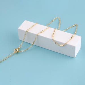May include: A delicate gold-colored necklace with a chain and small, cylindrical bar accents. The necklace is displayed on a white rectangular block against a light blue background. The necklace has an adjustable length.