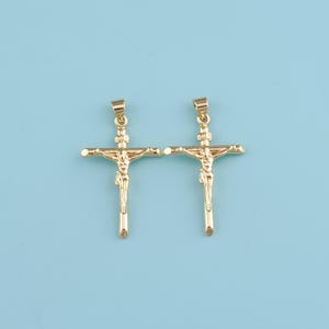 Puede incluir: Dos colgantes de crucifijo dorados, cada uno representando a Jesús en la cruz. Los colgantes están detallados con una figura de Jesús y están colocados sobre un fondo azul claro. Los colgantes están diseñados con un lazo en la parte superior para una cadena.