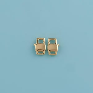 May include: Two gold-colored metal clasps, each with a rectangular body and rounded edges, are displayed against a light blue background. The clasps have a simple, functional design, with a small opening in the center.