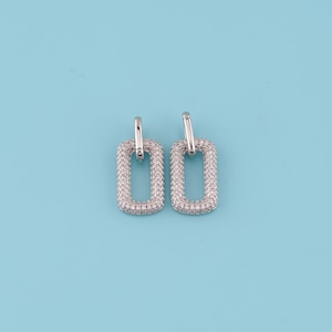 May include: A pair of silver-toned rectangular hoop earrings. Each earring features a rectangular frame encrusted with small, clear crystals. The earrings are set against a light blue background.