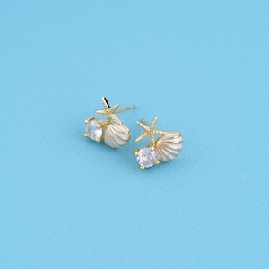 May include: A pair of gold stud earrings featuring a small starfish, a white seashell, and a clear round gemstone. The earrings are designed with a post back and showcase a nautical theme with metallic gold accents and iridescent white shell details.