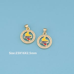 Op de afbeelding: Twee goudkleurige hangers met een rond ontwerp met de kaart van Colombia in geel, blauw en rood. De hangers zijn omgeven door kleine, heldere kristallen. De tekst "COLOMBIA" is zichtbaar. Afmetingen: 23x16x2,5 mm.