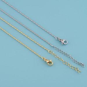 May include: Close-up of three delicate necklace chains in gold and silver tones. The chains have a textured design and are displayed against a light blue background. Each chain features a clasp and an extender chain.