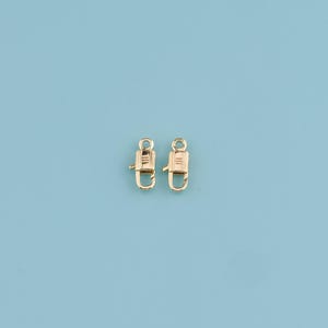 May include: Two gold-colored swivel clasps with a D-ring design. The clasps are positioned against a light blue background. These clasps are likely used for attaching straps or chains to bags, jewelry, or other accessories.