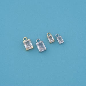 May include: Four rectangular clear gemstone charms with metal settings. Two charms feature gold-toned metal prongs and jump rings, while the other two charms feature silver-toned metal prongs and jump rings. Each charm has a faceted surface.