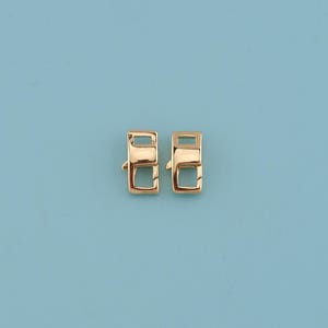 May include: Two gold-colored rectangular clasps with a rounded top and a small hook on the side. The clasps are set against a light blue background. These clasps are likely used for jewelry making or other craft projects.