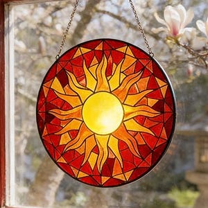 Puede incluir: Un atrapasoles redondo de vidrieras con un diseño de sol en tonos rojos, naranjas y amarillos cuelga de una ventana. El atrapasoles está suspendido por una cadena. Una pequeña foto enmarcada y una planta suculenta están en el alféizar.