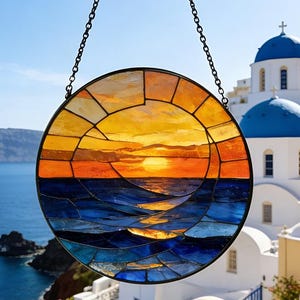 Puede incluir: Una vidriera redonda que representa una puesta de sol sobre el océano. La ventana está suspendida por una cadena y presenta tonos de naranja, amarillo y azul. El fondo muestra una escena costera con edificios blancos y cúpulas azules.