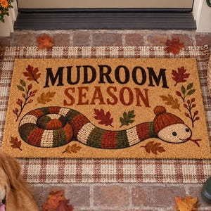 May include: A rectangular coir doormat with a tan background featuring the text MUDROOM SEASON in black and brown letters. The design includes a long, striped snake wearing a knit cap and scattered autumn leaves in shades of red, orange, and green.