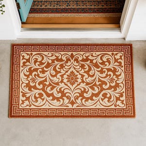 May include: A rectangular doormat with a floral design in shades of brown and cream. The mat features a border with a Greek key pattern. The doormat is placed in front of a doorway, with hanging plants on either side.