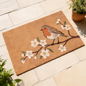 Può includere: Zerbino rettangolare con un'illustrazione dettagliata di un pettirosso posato su un ramo fiorito. L'uccello ha il petto rosso, ali marroni e testa grigia. Fiori bianchi con foglie verdi adornano il ramo su sfondo beige.