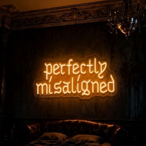 Op de afbeelding: Een oranje neonbord met de tekst "perfectly misaligned". Het bord is bevestigd aan een donkere muur boven een leren bank. Op de achtergrond is een kroonluchter te zien.
