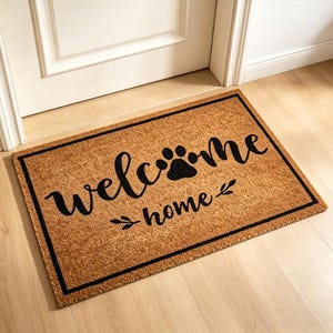 Könnte beinhalten: Rechteckige Fußmatte mit brauner Kokosoberfläche und schwarzem Rand. Die Matte zeigt die Worte "welcome home" in Schreibschrift, mit einem Pfotenabdruck und Blättern.