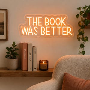 以下が含まれることがあります： 温かいオレンジ色の光で「THE BOOK WAS BETTER」と書かれたネオンサイン。その下に、本、火の灯ったキャンドル、鉢植えの植物が置かれた木製の棚があります。ベージュのアームチェアと錆色のクッションが前景にあります。