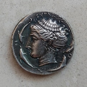 Puede incluir: Una moneda de plata que muestra el perfil de una mujer con cabello detallado y un tocado. Dos delfines flanquean la cabeza de la mujer. La moneda tiene una apariencia antigua y desgastada.