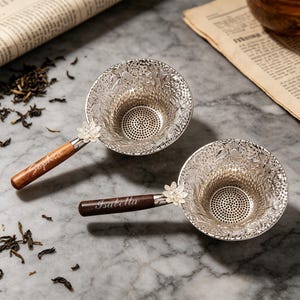 May include: Two silver tea infusers with floral designs and wooden handles. The handles are engraved with the names "Sophia" and "Isabella." Each infuser has a small white flower detail where the handle meets the strainer.