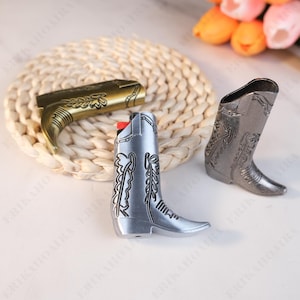 May include: Three cowboy boot-shaped lighters in gold, silver, and gunmetal grey. The silver lighter has a red ignition button. Each lighter features decorative embossed detailing, with the gold one lying horizontally.