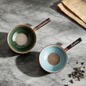 以下が含まれることがあります： 木製の持ち手が付いた陶器製の茶こし2点。1点はダークグリーン、もう1点はライトブルーです。持ち手にはそれぞれ筆記体で名前が刻印されており、緑の茶こしには「Charlotte」、青の茶こしには「Sophia」とあります。どちらも金属製の細かいメッシュフィルターが付いています。