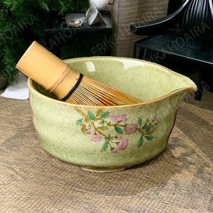 以下が含まれることがあります： 花柄の緑色の陶器製の抹茶碗と、中に置かれた竹製の茶筅。碗は注ぎ口と広い縁があります。