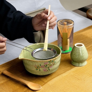 以下が含まれることがあります： 花柄の緑色の陶器のボウル、ステンレス製のストレーナー、透明なプラスチック容器に入った木製ホイスク、緑色の陶器のティーカップ、木製のスプーン。