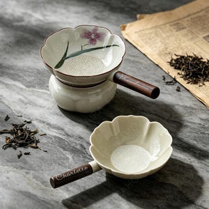 以下が含まれることがあります： 花形のボウルと木製の持ち手が付いた陶器製の茶こし2点。片方には紫色の花のイラストと持ち手にTheodoreの名前が記されています。もう片方はクリーム色で、持ち手にCharlotteの名前が記されています。
