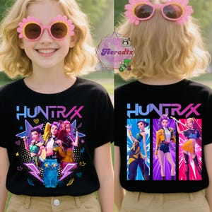 May include: Black t-shirt with the word "HUNTRAX" in blue and pink, featuring a graphic of a group of animated figures. The front design includes a blue cat and stars. The back design shows three figures in colorful outfits.