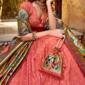 Puede incluir: Un atuendo tradicional indio de color coral con detalles dorados. El atuendo incluye una blusa con mangas estampadas, una falda y un bolso a juego con un diseño floral. El atuendo está adornado con joyas y pulseras doradas.