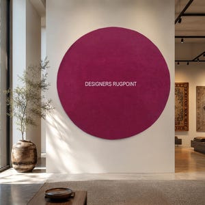 May include: A large circular magenta rug mounted on a wall, featuring the white text DESIGNERS RUGPOINT centered on its surface. The rug has a solid, uniform color and a flat, circular shape, displayed prominently against a neutral wall.