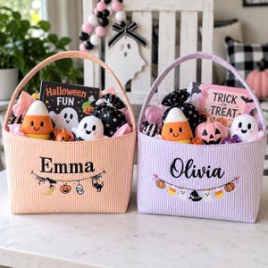 Puede incluir: Dos cestas de tela con asas, una naranja y otra lavanda, con los nombres bordados Emma y Olivia. Cada cesta contiene peluches que incluyen un fantasma y un caramelo de ma&iacute;z, una peque&ntilde;a calabaza y un libro titulado Halloween Fun o Trick or Treat.