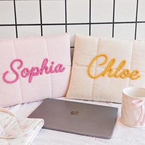 May include: Two rectangular quilted pillows, one light pink with the name Sophia in pink script and one cream with the name Chloe in yellow script. A grey laptop sits in front of the pillows next to a white mug featuring a pink bow detail.