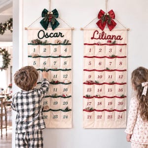 May include: Two off-white fabric wall-hanging advent calendars with twenty-four pockets each. One features green trim and the name Oscar, the other red trim and the name Liliana. Both are topped with a decorative bow and hanging greenery.