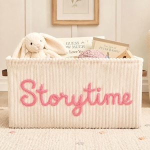 May include: A rectangular cream-colored corduroy storage bin featuring the word Storytime embroidered in pink yarn on the front. The bin is filled with a plush white rabbit toy, a pink crocheted item, and books titled Guess How Much I Love You.