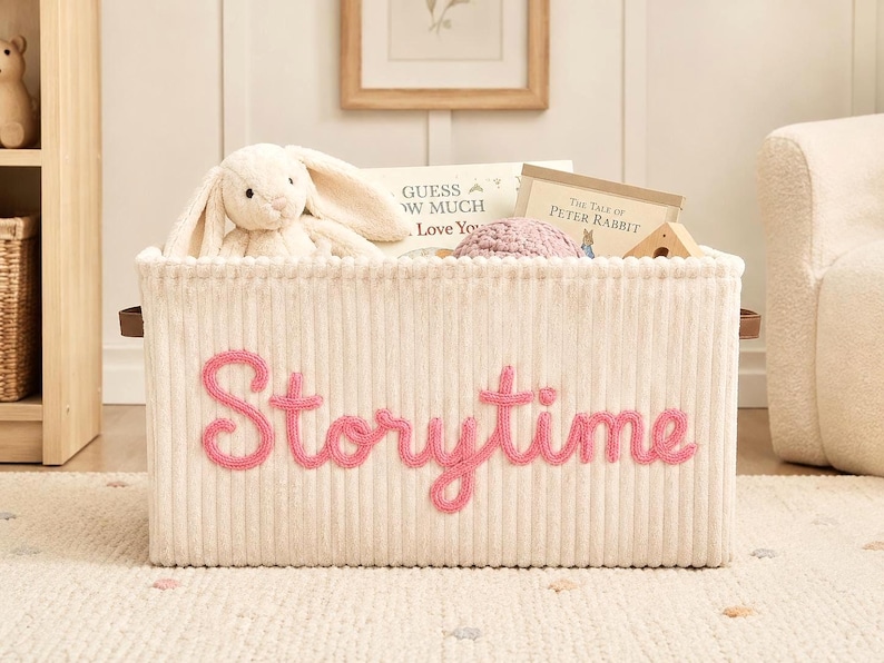 May include: A rectangular cream-colored corduroy storage bin featuring the word Storytime embroidered in pink yarn on the front. The bin is filled with a plush white rabbit toy, a pink crocheted item, and books titled Guess How Much I Love You.