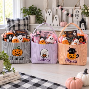 Puede incluir: Tres cestas de tela con asas, a rayas negras, lavanda y naranjas. Cada una presenta dise&ntilde;os bordados: una calabaza con el nombre Hunter, un fantasma con el nombre Lainey y un gato negro sobre una calabaza con el nombre Olivia. Las cestas contienen libros de actividades, vasos, peluches y dulces de Halloween.