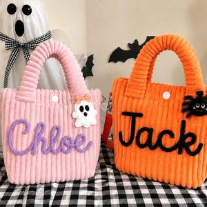 Puede incluir: Dos bolsos de mano de pana con asas superiores y correas para el hombro. El bolso rosa presenta el nombre Chloe en letra morada y un parche de fantasma. El bolso naranja presenta el nombre Jack en letra negra y un parche de araña.