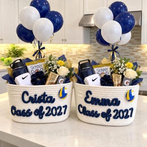 May include: Two white rope baskets with navy blue lettering reading Crista Class of 2027 and Emma Class of 2027. Each basket contains a navy water bottle, white Nike socks, a navy scrunchie, SkinnyPop, a flower, and a volleyball patch.