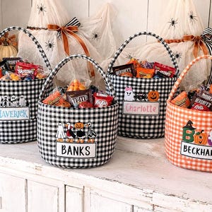 May include: Four fabric baskets with handles, three in black and white gingham and one in orange and white gingham. Each basket features embroidered Halloween motifs and a rectangular name patch with the names Maverick, Banks, Charlotte, and Beckham.
