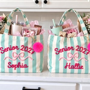 May include: Two canvas tote bags with vertical teal and white stripes. Each bag features pink embroidered text reading Senior 2027, a pink bow, and a name, Sophia or Hallie. A bright pink pom-pom keychain is attached to each bag handle.
