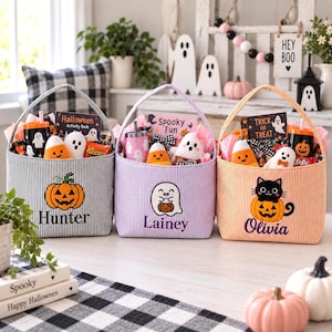 Puede incluir: Tres cestas de Halloween de tela con asas, con rayas verticales en gris, morado claro y naranja. Cada cesta está bordada con un nombre: Hunter, Lainey u Olivia, y un personaje: una calabaza, un fantasma o un gato negro.