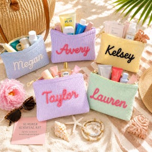 May include: Five rectangular corduroy zipper pouches in light blue, lavender, yellow, purple, and mint green, each embroidered with a name: Megan, Avery, Kelsey, Taylor, and Lauren. The pouches are filled with various skincare and beauty products.