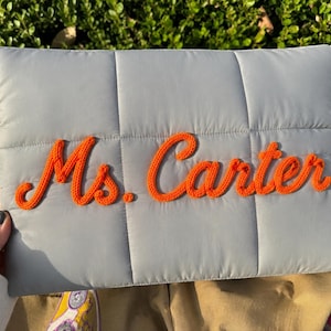 May include: A rectangular light gray quilted pouch featuring the name Ms. Carter embroidered in bright orange yarn across the center. The surface has a grid-like stitched pattern, and the text is rendered in a cursive, raised script style.