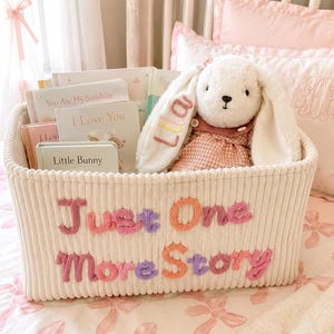 May include: A cream-colored corduroy storage basket featuring the embroidered text Just One More Story in pink, purple, and orange. Inside the basket are several children's books titled You Are My Sunshine, I Love You, and Little Bunny, plus a plush white bunny toy with the name Lila embroidered on its ear.