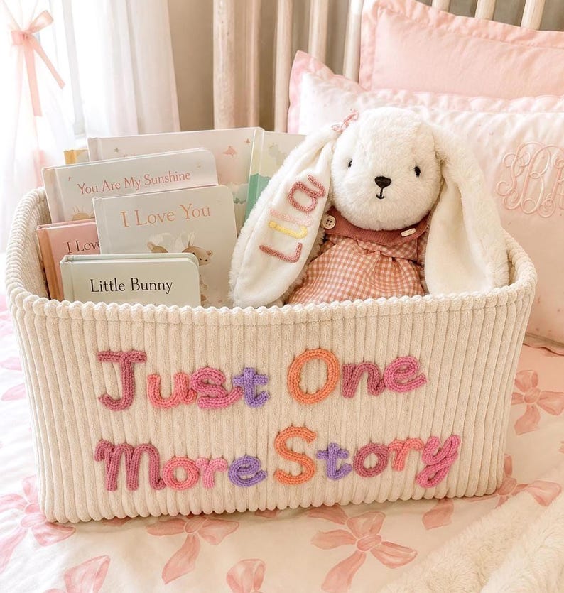 May include: A cream-colored corduroy storage basket featuring the embroidered text Just One More Story in pink, purple, and orange. Inside the basket are several children's books titled You Are My Sunshine, I Love You, and Little Bunny, plus a plush white bunny toy with the name Lila embroidered on its ear.