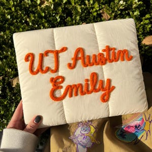 May include: A rectangular, off-white, quilted pouch featuring the orange, textured, embroidered text "UT Austin" on the top line and "Emily" on the bottom line. The fabric has a smooth, padded appearance with vertical stitching lines.
