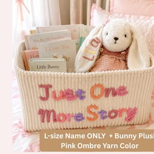 May include: A cream-colored corduroy storage basket with the text Just One More Story embroidered in pink, purple, and orange yarn. Inside are several children's books and a white plush bunny with the name Lia embroidered on its ear.
