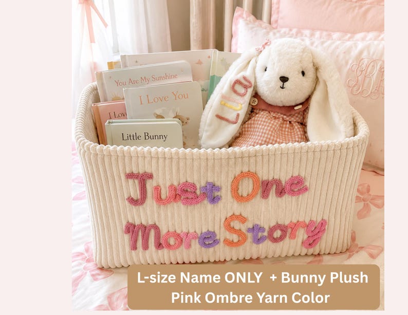 May include: A cream-colored corduroy storage basket with the text Just One More Story embroidered in pink, purple, and orange yarn. Inside are several children's books and a white plush bunny with the name Lia embroidered on its ear.