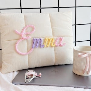 May include: A cream-colored quilted pillow featuring the name Emma in raised, multi-colored yarn lettering. Beside it sits a white ceramic mug with a pink bow handle and a closed gray laptop with a coiled white charging cable on top.
