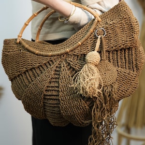 Puede incluir: Un gran bolso de mano de crochet marrón con un asa de bambú y un largo borla marrón. El bolso está hecho de un hilo grueso y texturizado y tiene una forma única y orgánica.