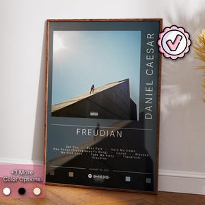 May include: A black and white poster with a brown wood frame. The poster features a photo of a person walking on a concrete structure. The text "FREUDIAN" is in large white letters at the bottom of the poster. The text "DANIEL CAESAR" is in white letters on the right side of the poster. The text "AUGUST 25, 2017" is in small white letters at the bottom of the poster.