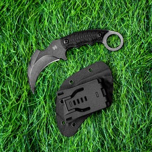 Puede incluir: Un cuchillo karambit negro con hoja curva y mango texturizado que presenta un anillo para el dedo en el extremo. El cuchillo incluye una funda negra con un clip para cinturón, mostrando un pequeño logotipo blanco en la hoja.
